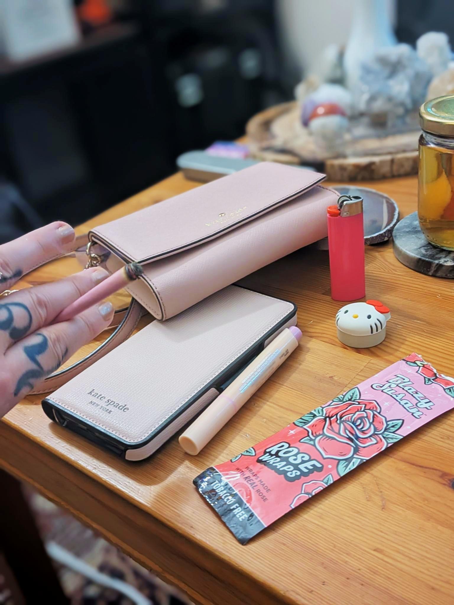 Picture of a table showcasing my pink Kate Spade wallet and phone case, my pink blunt as well as the packaging for the rose blunt wrap, my lip gloss, a pink lighter, and a Hello Kitty dab container.